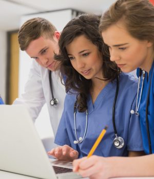 Grupo de estudantes de medicina com jalecos e estetoscópios, reunidos em volta de um notebook, concentrados em um trabalho colaborativo.