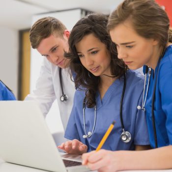 Grupo de estudantes de medicina com jalecos e estetoscópios, reunidos em volta de um notebook, concentrados em um trabalho colaborativo.