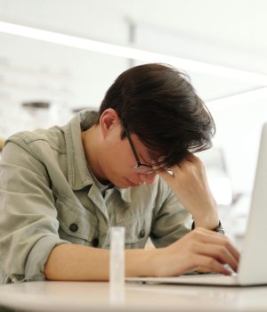 Jovem com expressão de cansaço e sinais de burnout, apoiando a cabeça enquanto trabalha no notebook em ambiente interno.