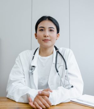 Médica jovem com jaleco e estetoscópio sentada à mesa em consultório, olhando para a câmera com expressão serena e profissional.