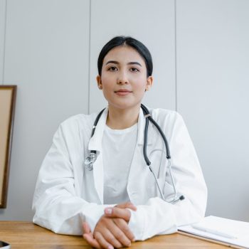 Médica jovem com jaleco e estetoscópio sentada à mesa em consultório, olhando para a câmera com expressão serena e profissional.