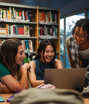 Três estudantes de Medicina estudam juntas em uma biblioteca, reunidas em torno de um notebook durante a semana de provas.
