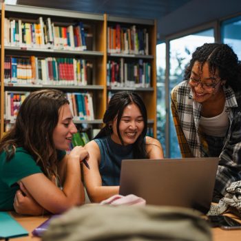 Três estudantes de Medicina estudam juntas em uma biblioteca, reunidas em torno de um notebook durante a semana de provas.