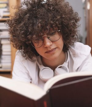 Pessoa de óculos lendo um livro de capa preta no chão, com fones de ouvido no pescoço.