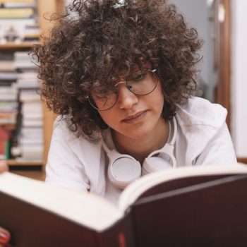 Pessoa de óculos lendo um livro de capa preta no chão, com fones de ouvido no pescoço.