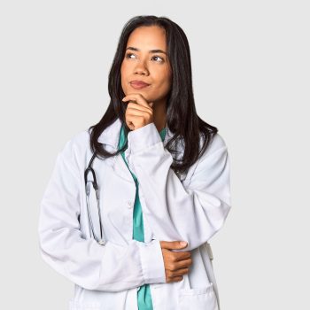 Mulher médica com expressão pensativa, vestindo jaleco branco e estetoscópio no pescoço, olhando para o lado.