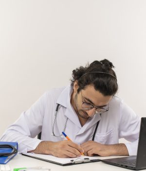 Médico jovem com jaleco e estetoscópio escreve em uma prancheta sobre uma mesa, ao lado de um notebook e itens médicos.