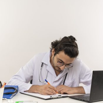 Médico jovem escrevendo em prancheta sobre a mesa com notebook e materiais de trabalho.