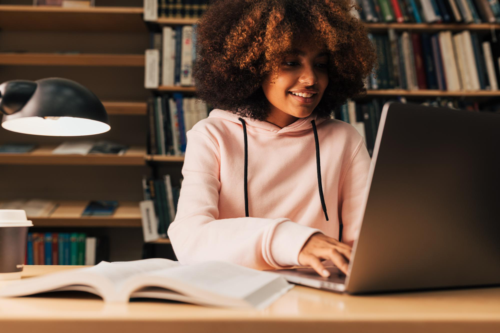 Uma jovem estudante, usando moletom rosa, está sentada em uma biblioteca estudando com o notebook aberto.