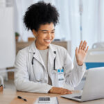 Médica sorrindo durante uma videochamada em um hospital, acenando para a câmera enquanto utiliza um notebook.