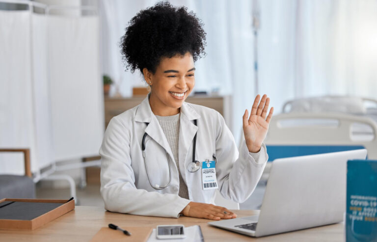 Médica sorrindo durante uma videochamada em um hospital, acenando para a câmera enquanto utiliza um notebook.