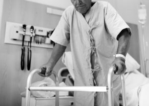 Idoso hospitalizado utilizando andador em quarto de hospital durante processo de recuperação física.
