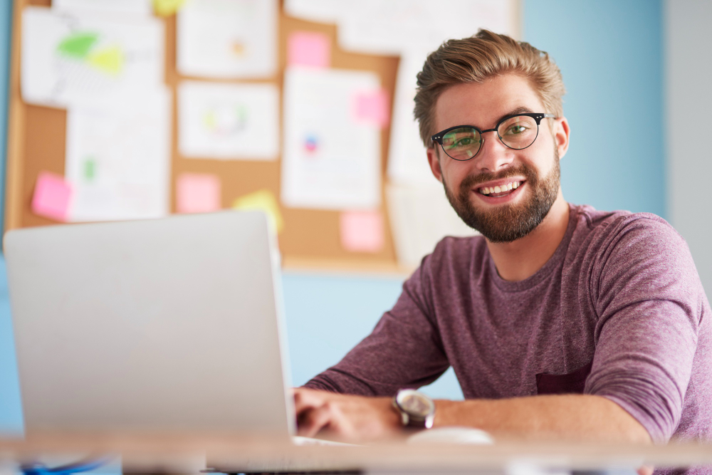 Homem jovem sorrindo enquanto trabalha em um laptop, com quadro de anotações ao fundo.