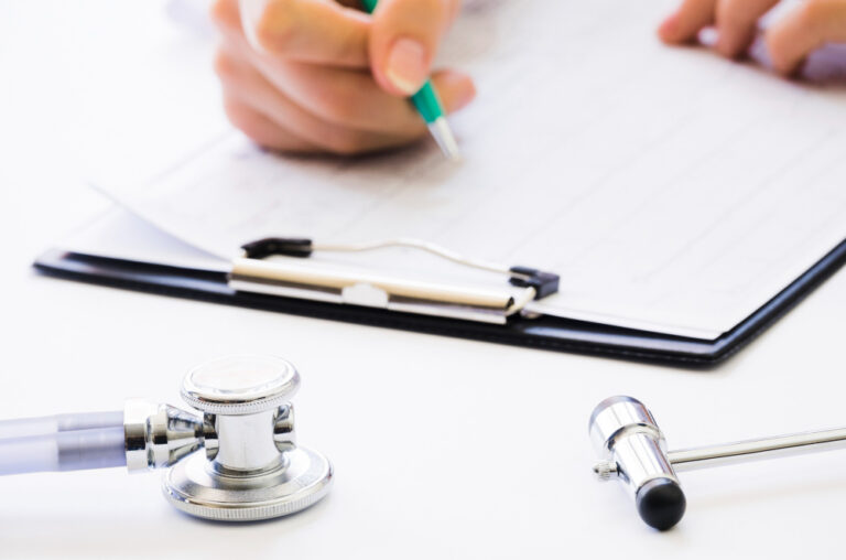 Estetoscópio e martelo neurológico sobre mesa médica enquanto profissional preenche formulário clínico em prancheta.