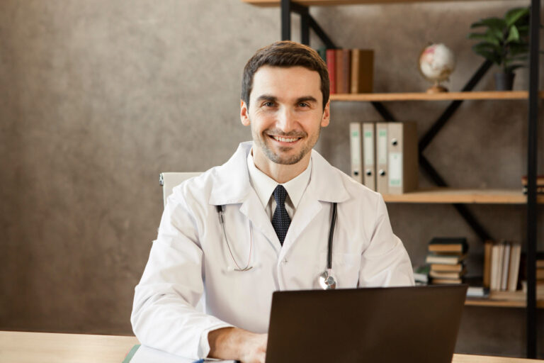 Profissional de saúde sorridente, usando jaleco branco e estetoscópio, sentado à mesa com notebook em consultório.