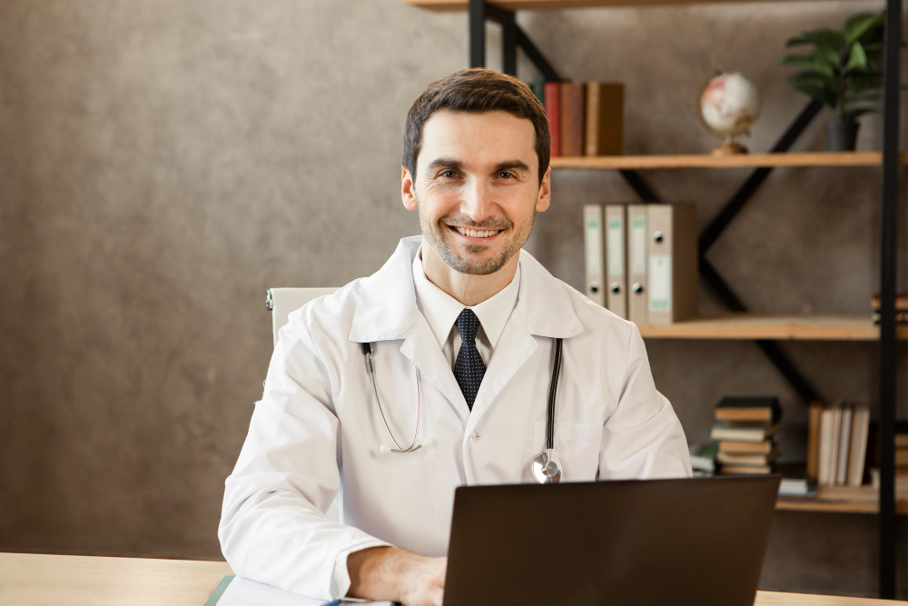 Profissional de saúde sorridente, usando jaleco branco e estetoscópio, sentado à mesa com notebook em consultório.