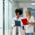 Duas profissionais da saúde conversando em corredor de clínica ou hospital enquanto analisam pranchetas médicas.
