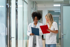 Duas profissionais da saúde conversando em corredor de clínica ou hospital enquanto analisam pranchetas médicas.