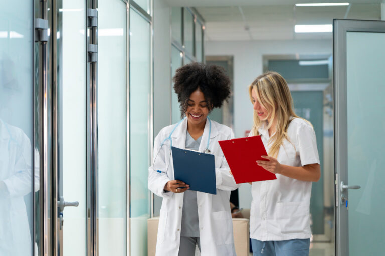 Duas profissionais da saúde conversando em corredor de clínica ou hospital enquanto analisam pranchetas médicas.