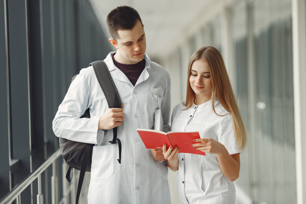 Fotografia de dois jovens estudantes de Medicina usando jaleco branco em um corredor iluminado. À direita, uma jovem loira segura um caderno aberto de capa vermelha e sorri enquanto lê. À esquerda, um rapaz com mochila nas costas observa as anotações ao lado dela.