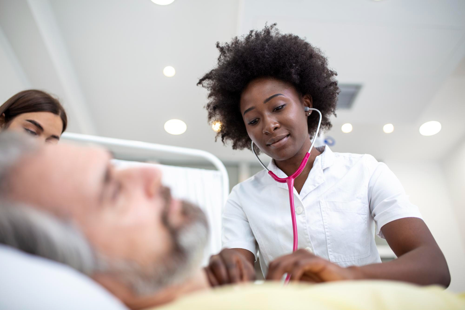 Médica negra, com cabelo black power, usando jaleco branco e estetoscópio rosa, examina um paciente idoso deitado em uma cama de hospital. Uma mulher mais jovem observa ao fundo, ao lado da cama.