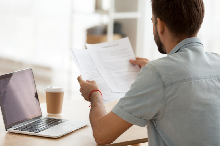 Homem analisando documentos em uma mesa de trabalho ao lado de um notebook.