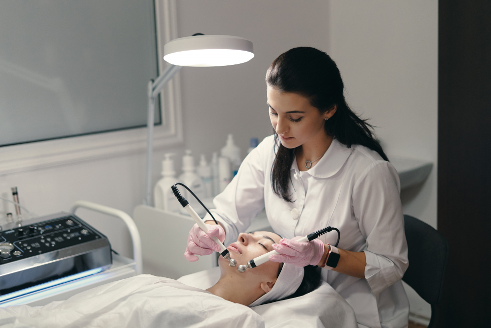 Biomédica esteta de jaleco branco e luvas rosas realiza procedimento estético facial com aparelho tecnológico em paciente.