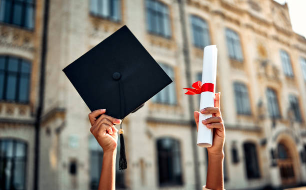 Mãos de uma pessoa negra erguendo um capelo de formatura e um diploma enrolado com laço vermelho em frente a um prédio universitário clássico.