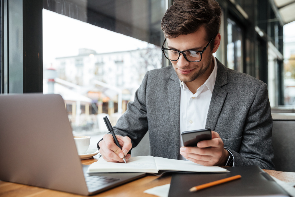 Homem jovem de óculos e blazer cinza, sorrindo enquanto escreve em um caderno e segura um celular, em uma mesa de trabalho com notebook aberto.