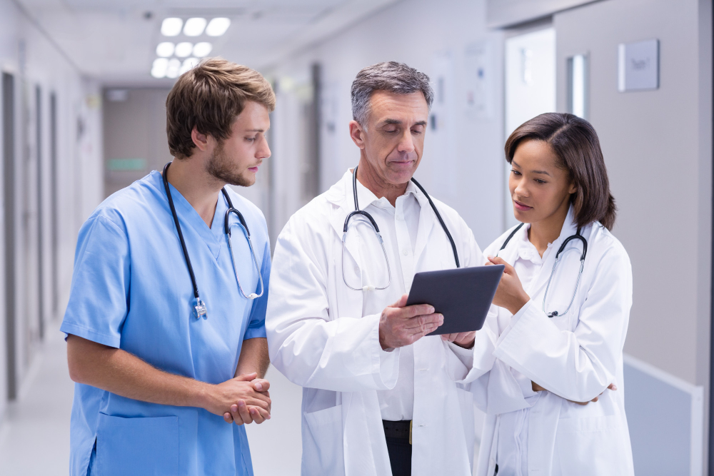 Médico e estudantes de medicina em um corredor hospitalar analisando dados em um tablet durante o internato.