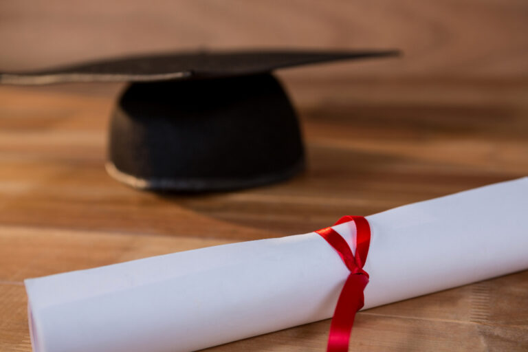 Diploma enrolado com fita vermelha ao lado de um capelo de formatura sobre uma mesa de madeira.