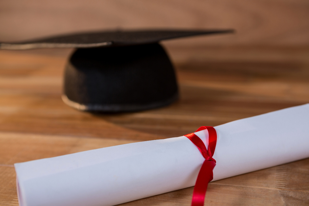 Diploma enrolado com fita vermelha ao lado de um capelo de formatura sobre uma mesa de madeira.