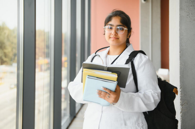 Estudante de Medicina com jaleco branco e livros nas mãos, em um corredor iluminado por janelas grandes, simbolizando o aprendizado e a rotina médica.