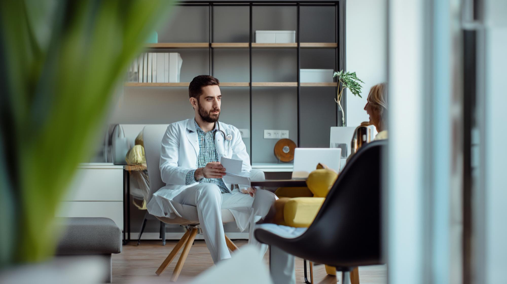Cena de uma consulta em um consultório moderno e bem iluminado. No centro, o nutricionista, um homem jovem de barba e jaleco branco, está sentado e conversa com atenção, segurando papéis. À sua frente, de costas para a câmera, está a paciente, uma mulher loira sentada. Ao fundo, prateleiras organizadas e uma janela com luz natural. No primeiro plano à esquerda, folhas verdes desfocadas emolduram a cena.