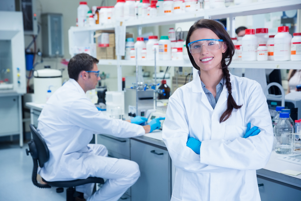 Cientista sorrindo em laboratório com colega ao fundo realizando experimentos.