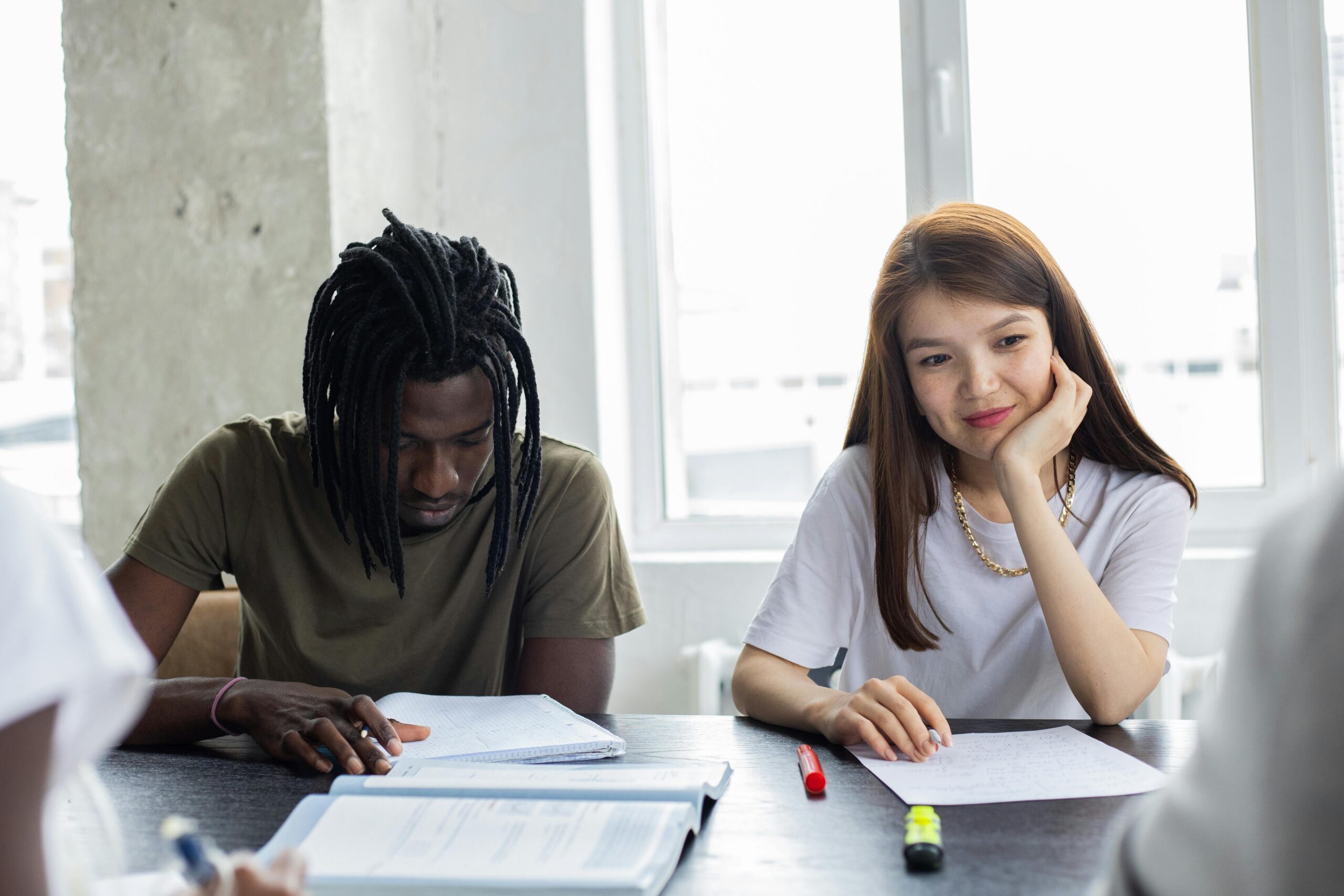 Dois estudantes sentados em uma mesa durante uma atividade em grupo, analisando anotações e materiais de estudo em uma sala iluminada.