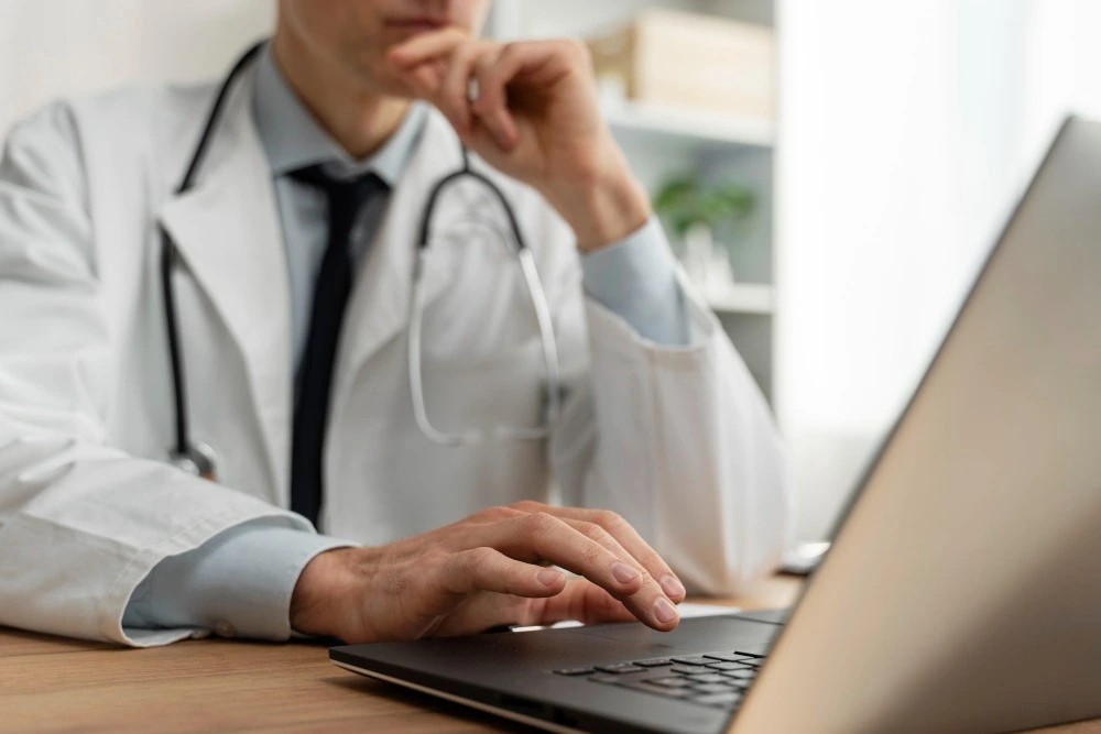 Médico de jaleco branco, gravata preta e estetoscópio utiliza um notebook, com uma das mãos apoiada no queixo, em ambiente de consultório.