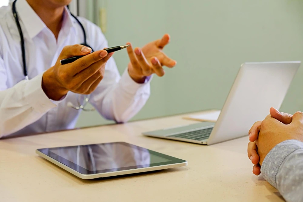 Médico com estetoscópio gesticula e segura uma caneta durante consulta com paciente, em mesa com notebook e tablet.