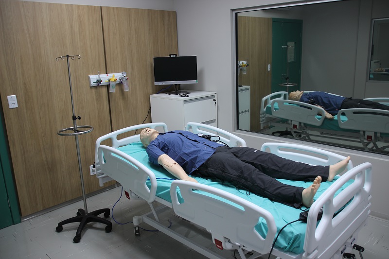 Boneco anatômico de simulação médica deitado em uma cama hospitalar dentro de uma sala equipada.