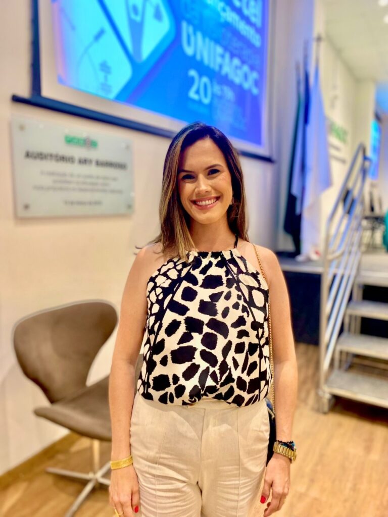 Mulher sorridente em pé em um auditório, vestindo blusa preta e branca estampada em padrão animal print e calça clara de alfaiataria, com um telão azul projetado ao fundo.