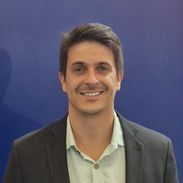 Homem sorridente de cabelos castanhos curtos, usando blazer escuro sobre camisa clara, posando diante de um fundo azul liso.