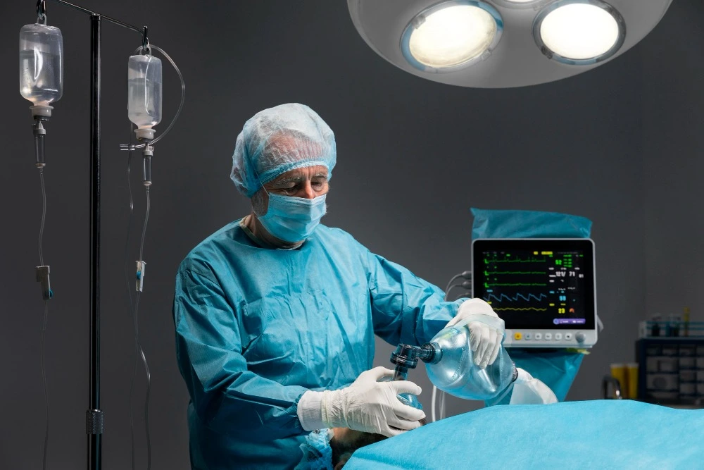 Profissional de saúde com roupa cirúrgica azul, máscara, touca e luvas realiza ventilação manual em paciente com auxílio de reanimador, em sala cirúrgica equipada com monitor de sinais vitais.