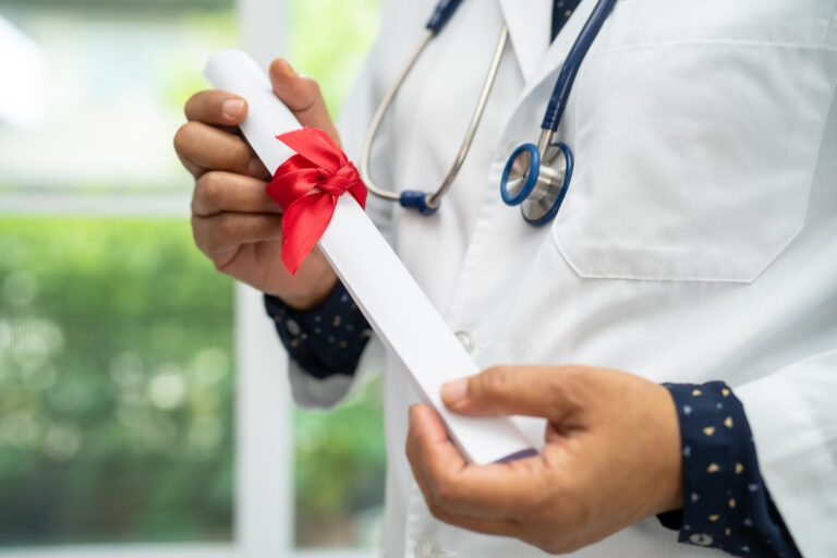 Profissional da saúde usando jaleco e estetoscópio segurando diploma enrolado com laço vermelho.