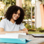 Jovem sorridente estuda ao ar livre, escrevendo em um caderno sobre a mesa com livros e laptop abertos.