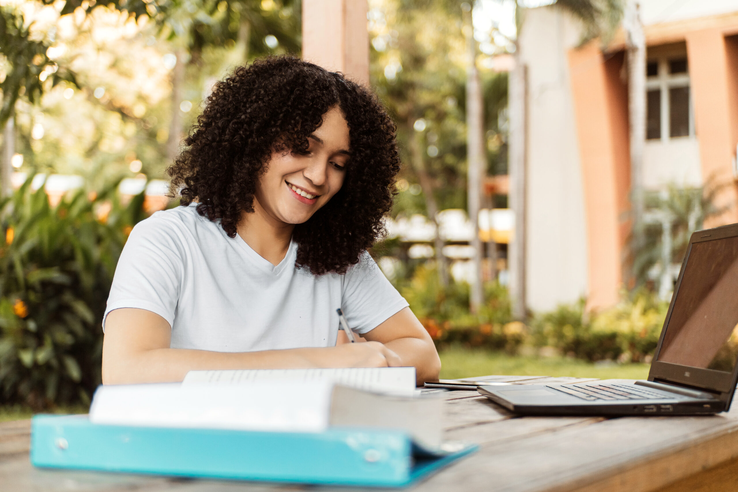 Jovem sorridente estuda ao ar livre, escrevendo em um caderno sobre a mesa com livros e laptop abertos.