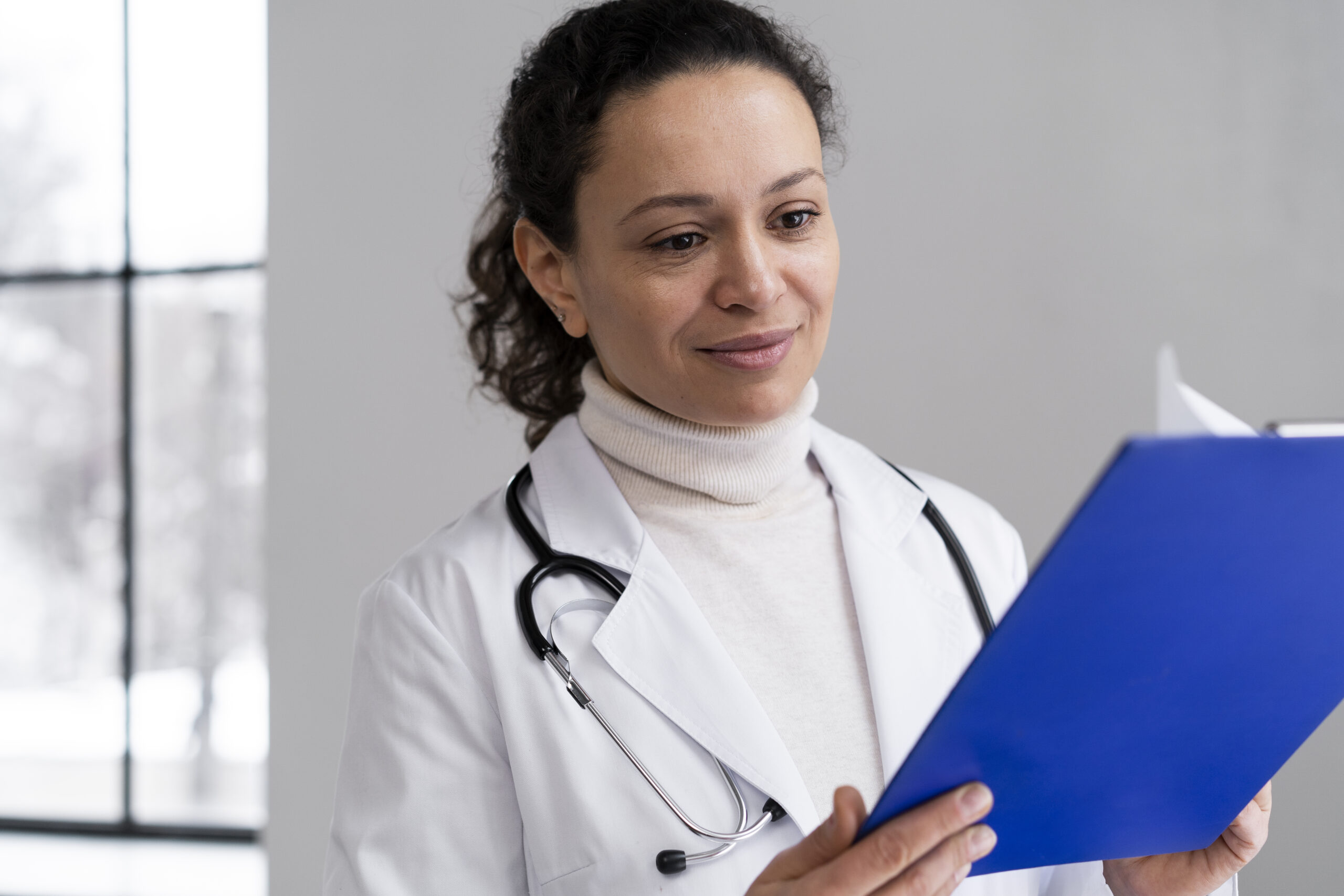 Médica com jaleco e estetoscópio olha atentamente para uma prancheta azul, em ambiente iluminado por luz natural.