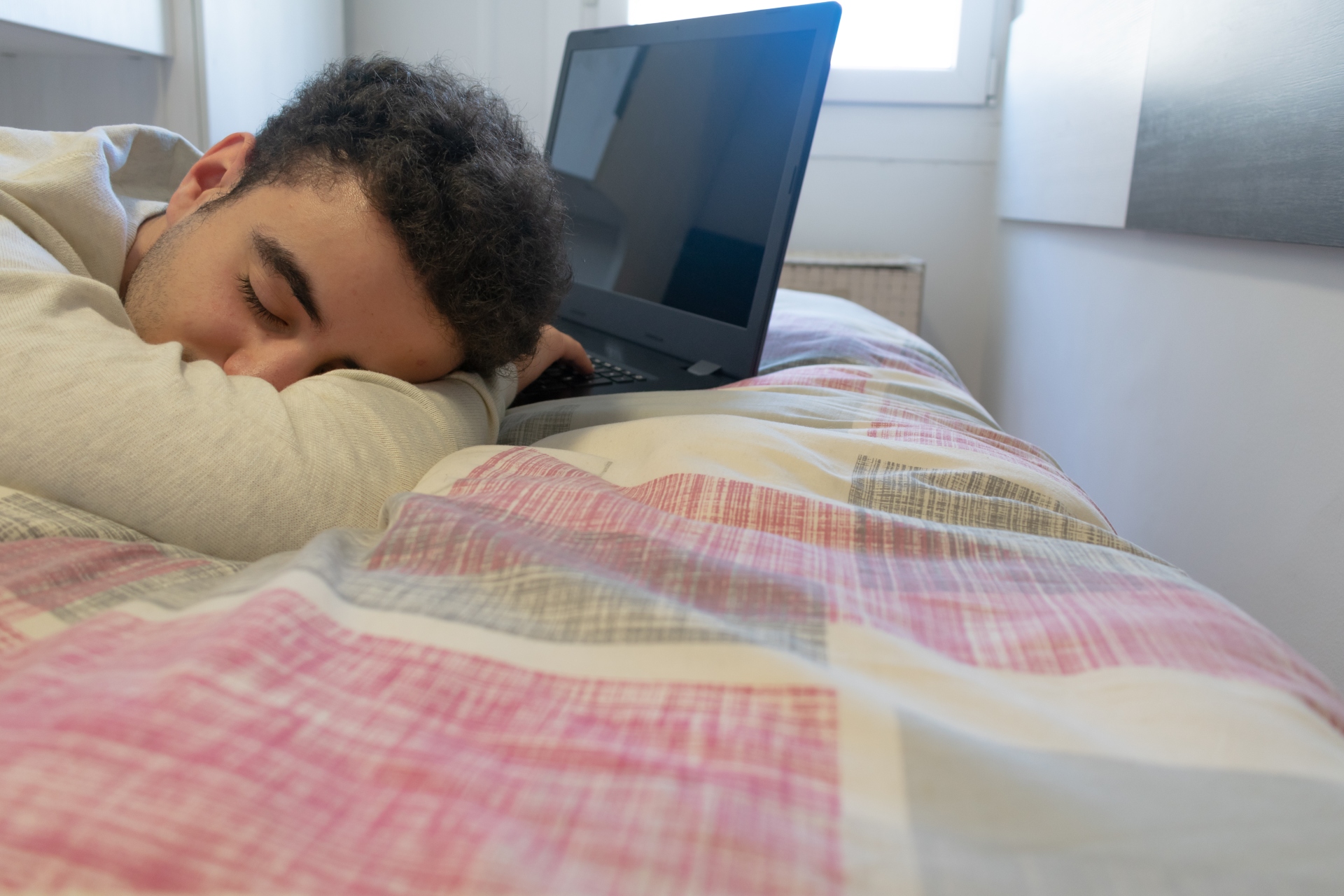 Jovem adormecido sobre a cama com um notebook aberto à frente, após trabalhar ou estudar por muito tempo.