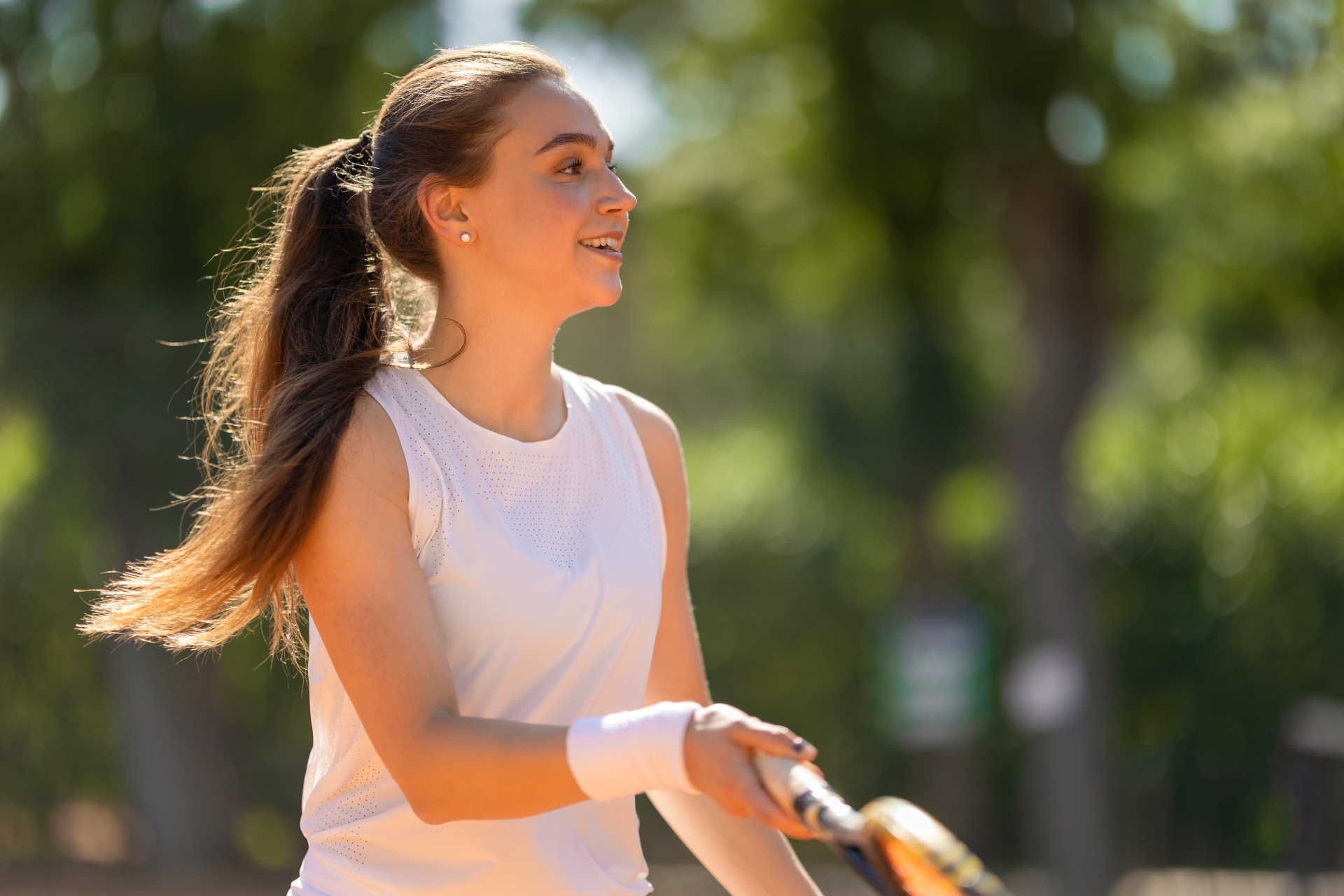 Jovem jogando tênis ao ar livre, segurando uma raquete e sorrindo durante o movimento, com árvores desfocadas ao fundo.