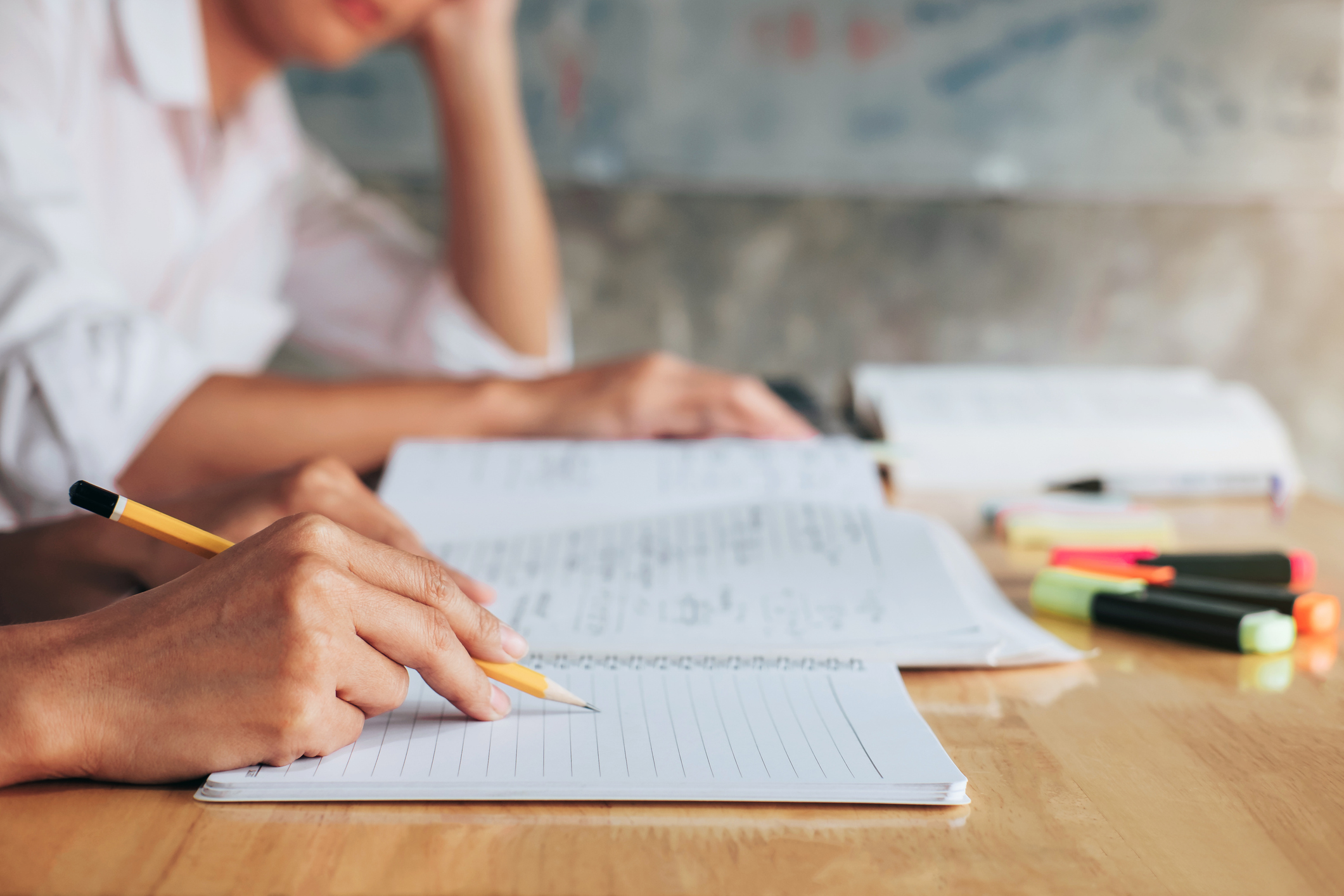 Pessoa escrevendo em um caderno com lápis enquanto estuda, com livros e marcadores de texto sobre a mesa