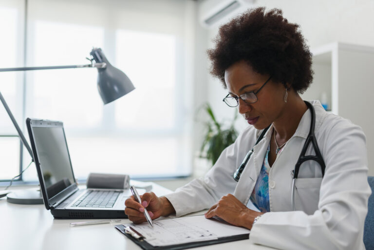 Uma médica usando um jaleco branco e óculos está sentada em uma mesa de escritório, escrevendo em uma prancheta. Há um notebook e uma luminária na mesa.
